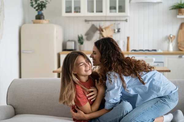 Happy caring mother hugs and tickles teenage girl to cheer up and give positive emotions during weekend or vacation. Joyful cheerful woman entertains school age daughter sits on sofa in apartment