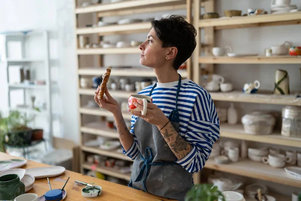 stock image Creative Spanish woman owner of pottery studio. Female artist in apron holding handcrafted clay mug with coffee, enjoying lunch in workshop, thinking, looking at window. Ceramics master at workplace