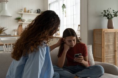 Sad teen girl with phone in hands does not want to communicate with mother ignoring parents and others. Emotionless schoolgirl sitting on sofa next to woman who is trying to make friends. Bullying. 