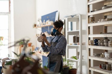 Self-sufficient woman in respirator works in crafts workshop grinding ceramic dishes after clay dried. Girl craftswoman is engaged in creative work earning on own hobby and selling handmade products