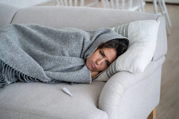 Sick woman with cold symptoms feeling unhappy and depressed, covered in blanket lying on sofa. Sad female suffering from fever with shivering and chills. Flu and depression concept