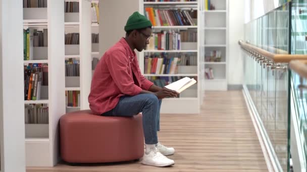Молодой Афроамериканец Читает Книги Современной Библиотеке Сидит Между Книжными Шкафами — стоковое видео