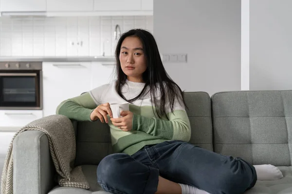 Portrait of young relaxed Asian woman sitting on sofa holding cup of tea or coffee enjoying morning daily ritual, looking at camera. Korean girl drinking hot warming winter drink while resting at home