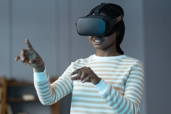 Focused african young woman wearing virtual reality glasses touching object with fingers, feeling excited while interacting with immersive digital world at home, selective focus. Cyberspace, vr game.