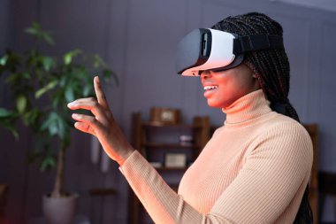 Excited african woman using VR video technologies raising hand up with mouth slightly opened while standing in red neon light at cozy modern office, black female shopping in virtual reality