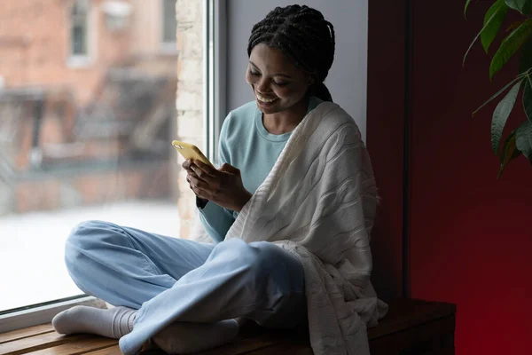 满脸笑容的黑人女人坐在窗台上 在网上商店里买东西 非洲裔美国年轻女性 带着温暖的毛毯在窗边和朋友们发短信 — 图库照片