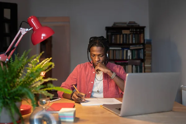 Pensive thoughtful young African American man with braids work on project pondering strategy make note solving problem analysing prepare to exam. Focused black guy student with laptop studying at home