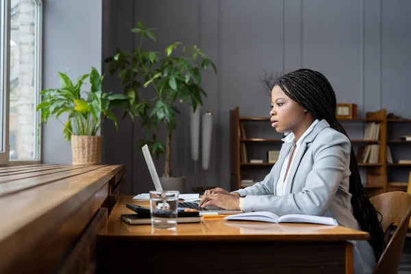 緑の観葉植物と居心地の良い自宅のオフィスに座っている間 ラップトップコンピュータ上の財務報告書を準備スーツリモートブックキーパーでアフリカ系アメリカ人女性に焦点を当て 選択的フォーカス リモート会計 — ストック写真