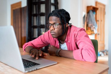 Unhappy African American guy remote worker sitting at home office, using laptop, doing boring freelance project, tired sad freelancer feeling uninspired and unmotivated. Freelancing and mental health