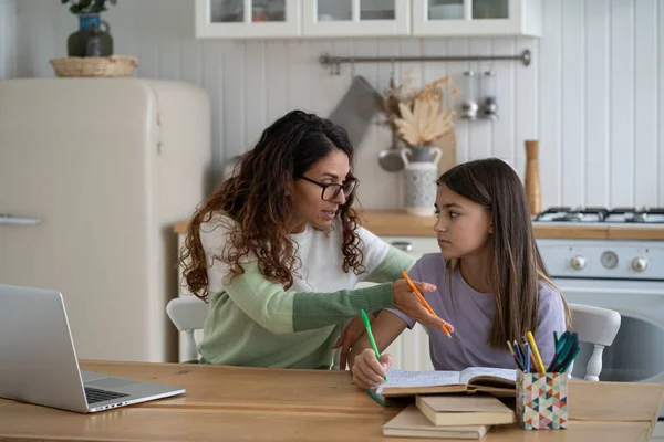 Nervous emotional mother helps daughter prepare report for school sits with textbook at kitchen table. Confused girl teenager reads books and makes notes in workbook under supervision of woman tutor