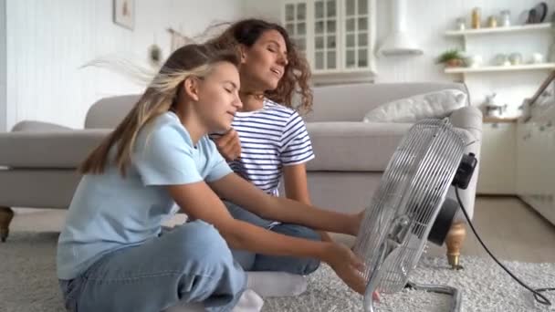 快乐的白人妇女和少女坐在客厅的地板上 享受着电扇吹来的风 年轻的母亲和乐观的女儿在炎热的天气里呆在家里享受凉爽的夏日 — 图库视频影像