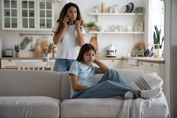 Disappointed parent mother talking on smartphone yelling at teenage daughter sitting on sofa, strict mom speaking with school teacher by phone scolding teen girl kid. Family conflict, teen discipline