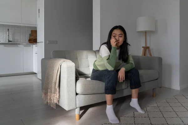 stock image Upset unhappy Asian woman sits on couch propping head with hand and looking into distance without initiative. Unmotivated sad Korean girl suffering from depression spending time alone in living room 