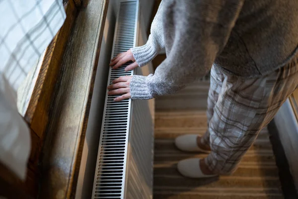 Closeup of woman in woolen sweater wants to keep warmth in apartment. Lady puts hands on room central heating battery to warm up and prevent illness because of coldness. Person checks work of heater
