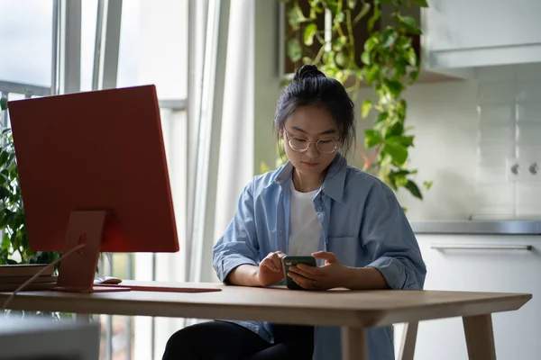 Asian woman sitting at remote workplace holding smartphone, using business application or mobile work tool, female freelancer checking email on mobile phone while working on computer at home office