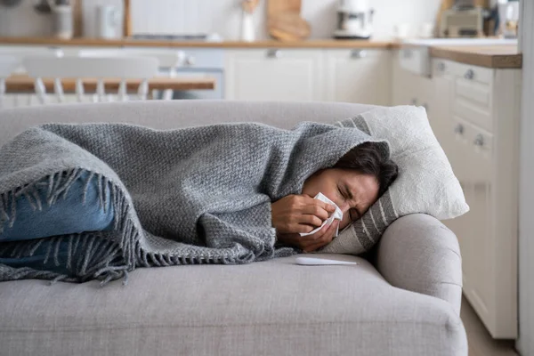Young woman lying under blanket on sofa coughing and sneezing, using paper tissue to blow her nose. Unhealthy female caught cold or flu, resting on couch, suffering coronavirus. Cold homes and health