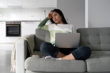 Thoughtful young Asian woman freelancer tired sits on sofa with laptop on lap suffering lack of motivation to work. Sad Chinese girl writer or journalist is having trouble with shortage inspiration