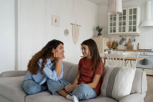 Young loving caring mother talking with teen girl daughter while sitting on sofa in living room at home, parent spending quality time with child. Trust between parents and children