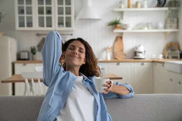 Happy relaxed woman enjoying lazy day at home, holding cup of tea and stretching body with closed eyes, resting on sofa in cozy apartment, taking break from daily routine, drinking morning coffee