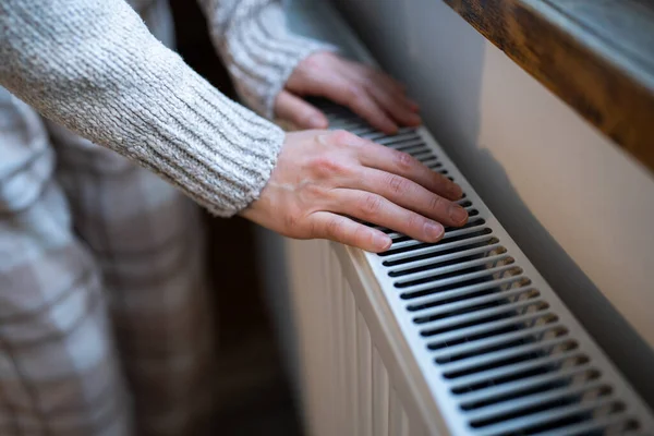 Closeup of woman in woolen sweater wants to keep warmth in apartment. Female puts hands on room central heating battery to warm up and prevent illness because of coldness. Person checks work of heater