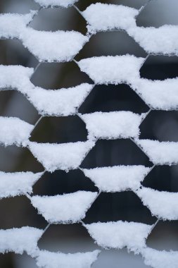 Street fence of thin mesh hawser with snowflakes accumulated in cells and half filled holes. Fragment of enclosure made of twisted wire forming hexagons filled with snow in cold winter weather clipart