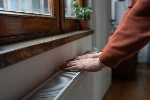 Closeup of man in sweater wants to keep warmth in apartment. Female puts hands on room central heating battery to warm up and prevent illness because of coldness. Person checks work of heater