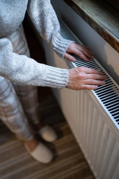 Closeup of woman in woolen sweater wants to keep warmth in apartment. Female puts hands on room central heating battery to warm up and prevent illness because of coldness. Person checks work of heater
