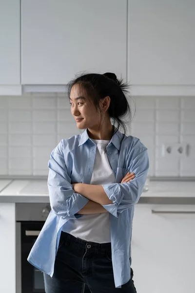 Happy Asian millennial woman wearing casual clothes standing in modern renovated kitchen at home, looking aside and smiling, enjoying weekend and free time. Satisfied female moved into new apartment
