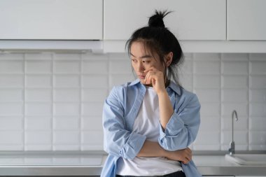 Unhappy depressed young Asian woman worrying about family problems, focusing on bad things. Frustrated upset female standing in kitchen at home. Domestic abuse and mental health, overthinking concept clipart