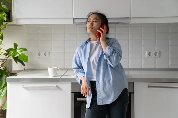 Worried confused millennial Asian girl having unpleasant conversation on mobile phone, dissatisfied Korean woman housewife holding smartphone waiting on hold while standing in kitchen at home