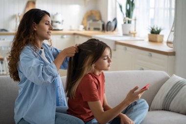 Young woman loving caring mother getting schoolkid ready for school in morning at home, braiding daughters hair while sitting together on sofa in living room. Childcare and parenthood concept