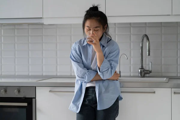 Stressed nervous young Asian woman standing in kitchen drinking water calming down after panic attack at home. Korean millennial girl feeling thirsty. Dehydration and anxiety