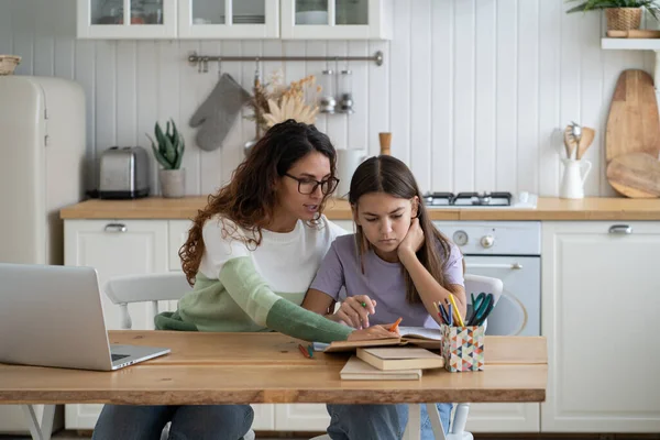Young mother teaching teen girl daughter at home, mom helping sad frustrated kid with difficult school task, supporting child in remote learning. Woman freelancer balancing parenting and remote work