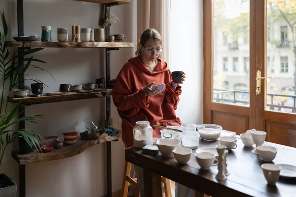Young woman potter resting in cozy home pottery studio, drinking tea and using smartphone. Female ceramist sitting at work table, taking break. Ceramic teacher having coffee break between lessons