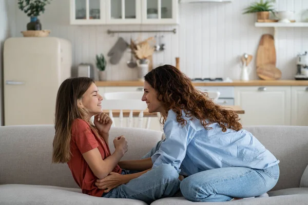 Positive single mother spending time with beloved child daughter at home, sitting on sofa at home and having fun. Parent mom tickling happy pre-teen girl, making kid laugh. Family leisure concept