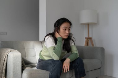 Pensive depressive Korean girl sitting on sofa looking into distance, blaming herself, thinking about failure. Anxious worried young Asian woman dealing with depression, having psychological problems