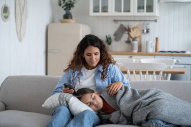 Loving caring mother woman sits on sofa in living room and smiles looking at teenager daughter falling asleep on knees. Happy kind casual girl nanny admires sleeping girl covered with warm blanket