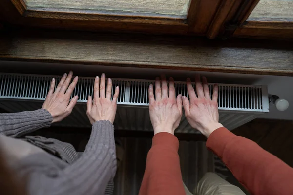 Man and woman warm hands by placing on radiator located by window of apartment due to sharp onset of cold weather. Top view of palms of guy and girl lying on centralized in-house heating system