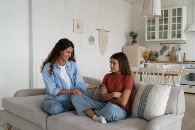 Cheerful caring woman trying to cheer up embarrassed school age daughter after house arrest. Shy introverted teenage girl sits in closed position on couch with mother wanting to play together