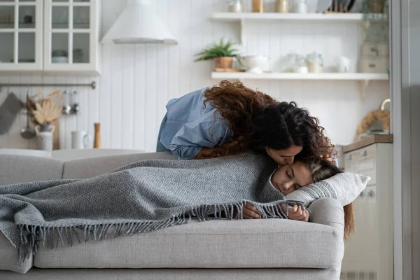 Young loving mother kissing daughter sleeping peacefully on couch during day nap time. Careful mommy giving kiss cute sleepy kid lying resting on sofa. Mom unconditional love, warmth and affection
