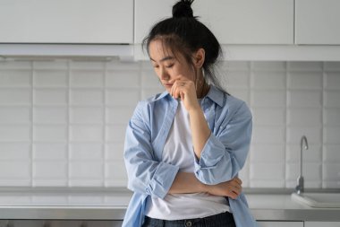 Offended embarrassed Asian woman looking down and feeling sad after breaking up with boyfriend. Discouraged depressed Korean girl standing in kitchen after stressful conversation with husband