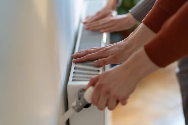 Closeup of man and woman warming hands on heaters hanging against wall on cold winter day. Owner of house changes temperature on radiator after start of heating season and arrival of first frost