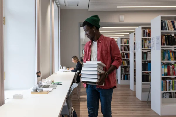 図書館やオンライン学習 アフリカ系アメリカ人の学生の男は 大学図書館で勉強しながら 本の山を保持し ノートパソコンの画面を見て 学習環境と教育の概念 — ストック写真
