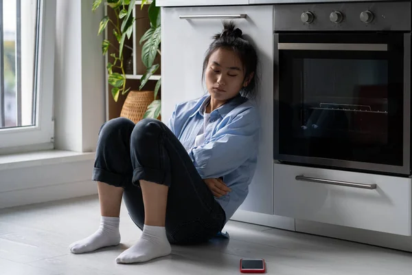 stock image Offended unhappy Asian woman sadly looking at phone sits on floor at kitchen. Sad thoughtful worried Korean girl experiencing depression after breaking up with beloved man or hurtful social media post