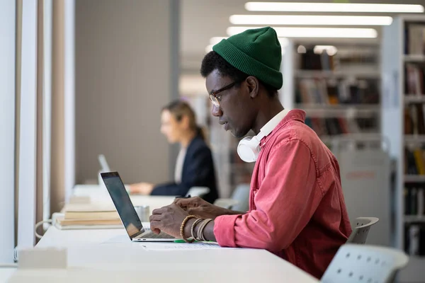 Entrepreneurial independent African American man sits with laptop in library building working on launching own IT startup. Concentrated casual guy students preparing report or essay for university