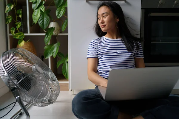 Relaxed carefree Asian woman sits on floor with laptop on lap and enjoying wind coming from fan. Optimistic Chinese girl blogger is typing article for website located near stove and house plants