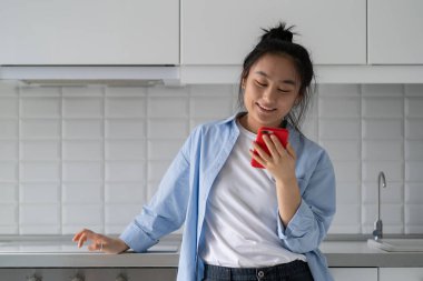 Happy millennial Asian girl holding smartphone reading positive uplifting news while spending time at home, positive young woman scrolling through social media, sending good morning message to friend