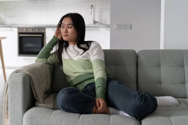 Carefree satisfied Asian woman enjoying loneliness and silence sits on sofa and remembering happy moments. Charismatic dreamy Japanese girl in casual clothes is waiting for evening to go on date