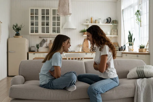 stock image Cursing nervous woman and teen girl are sits on couch and shouting out loud expressing resentment and feeling mutual dislike. Aggressive mom and daughter arguing not wanting to give in to each other