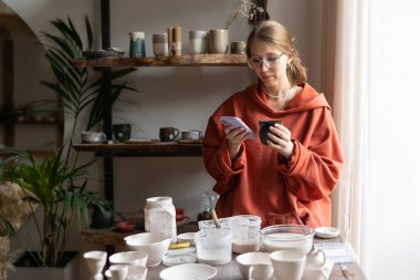 Focused woman seller ceramics shop standing in artist workshop with phone posting ads on social media. Craftswoman drinking coffee selling handmade utensils over internet, scrolling social networks